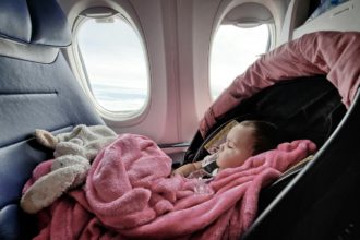 A baby girl is seen sleeping in her car seat while traveling on a plane wrapped in a pink blanket.