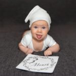 A cute baby is lying on a dark sofa and shows his tongue, rejoicing that he is already 3 months old