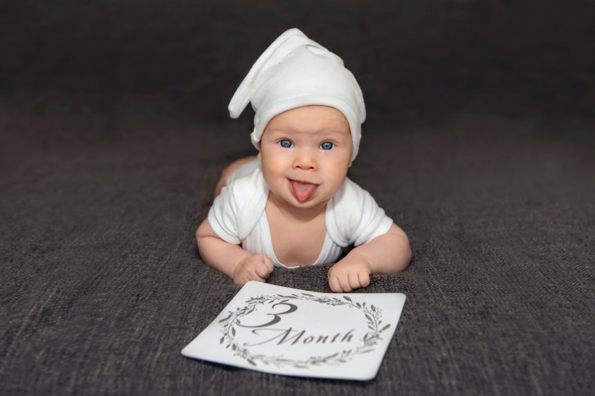 A cute baby is lying on a dark sofa and shows his tongue, rejoicing that he is already 3 months old